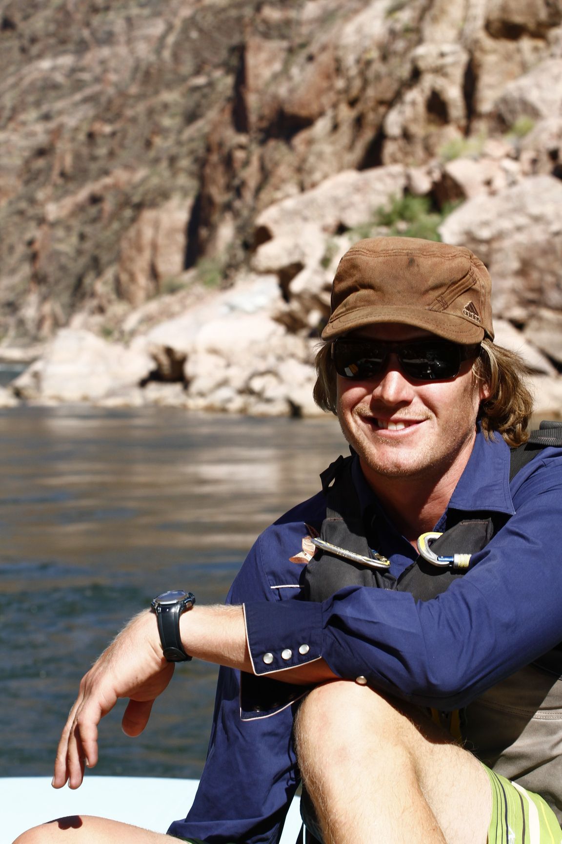 Man in hat and sunglasses smiles on a river, wearing a life vest with canyon in background.