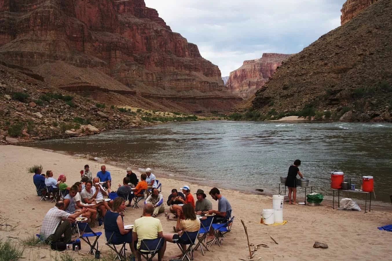 People seated in a circle on a sandy beach by a river in a canyon.