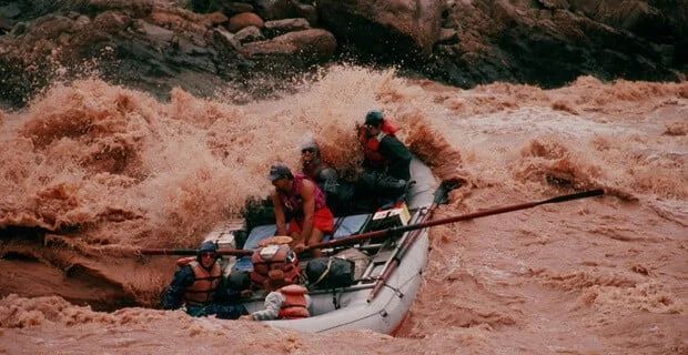 Raft navigating a raging, muddy river.  People in life vests battle waves. Brown water splashes.