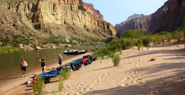 People and gear on a sandy riverbank in a canyon, near rafts.