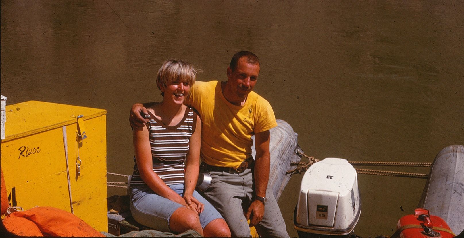 Man with arm around woman on a boat. Both smile, next to yellow box, motor, and river.