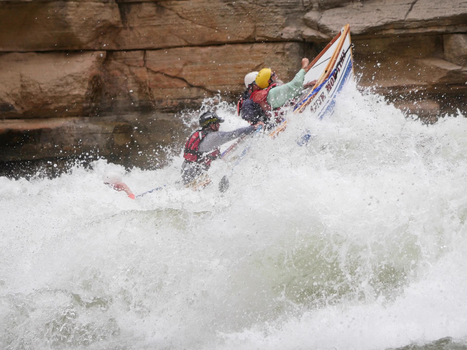 Whitewater rafters in a boat being tossed by a large wave. Rocky canyon walls in background.