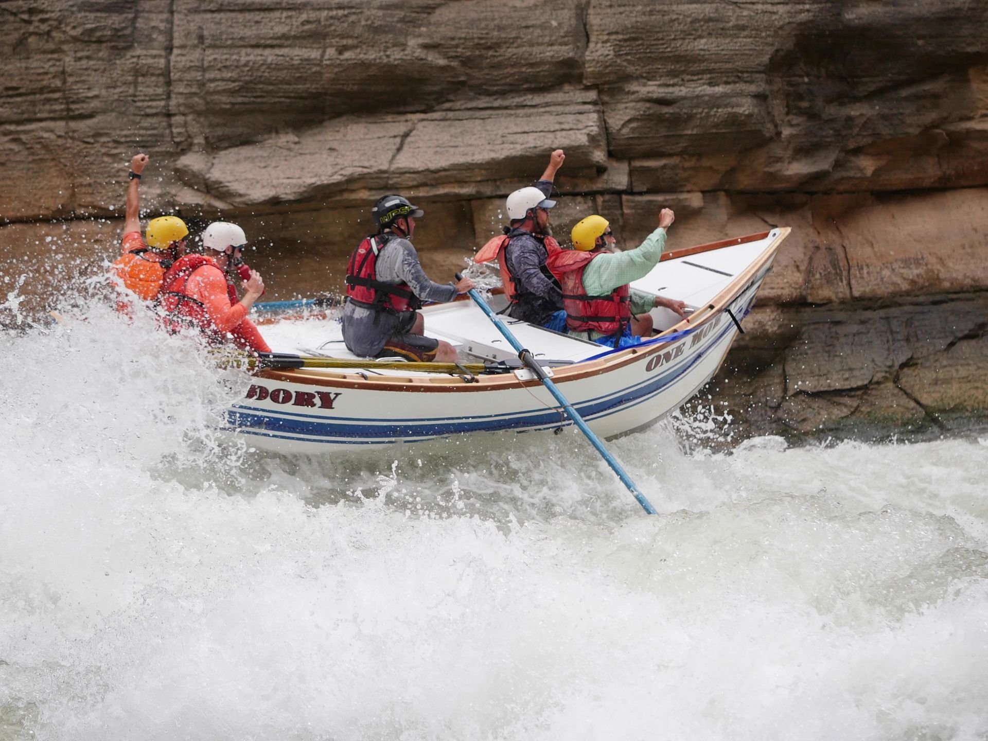 Whitewater rafting boat navigating rapids; passengers cheer, wearing life vests and helmets.