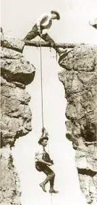 Man suspended by rope, dangling between cliffs while another man stands above, holding the rope.