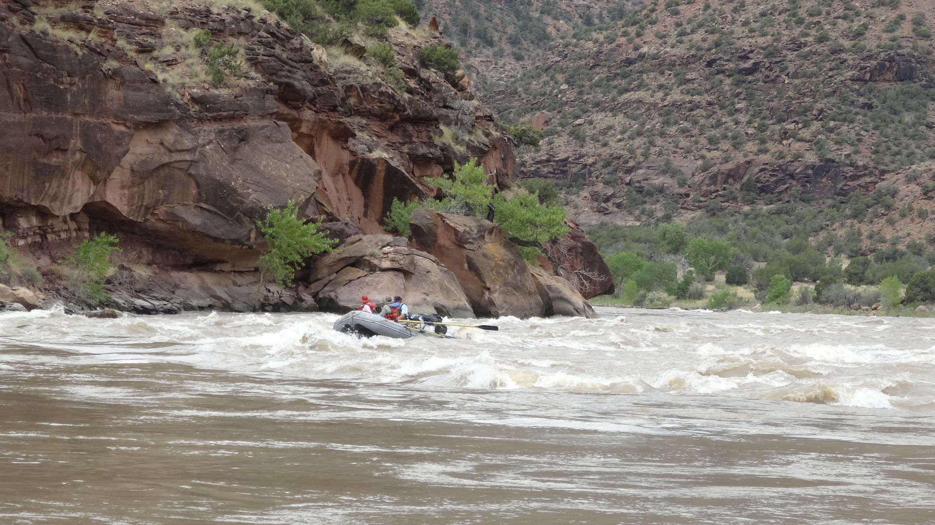 Whitewater raft on a churning river near a rocky cliffside, with several people visible.