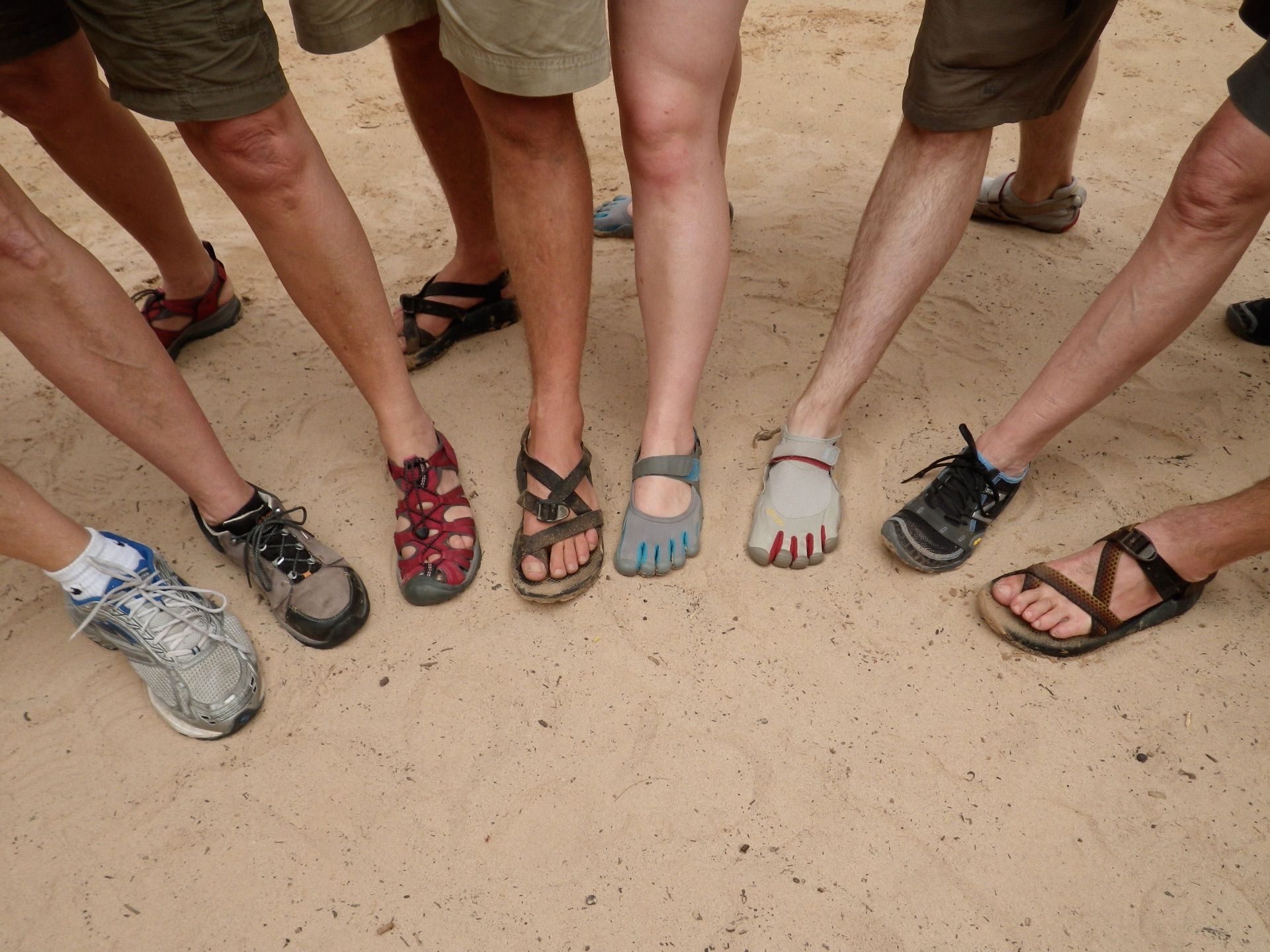 People's feet in various sandals and shoes standing on a sandy surface.