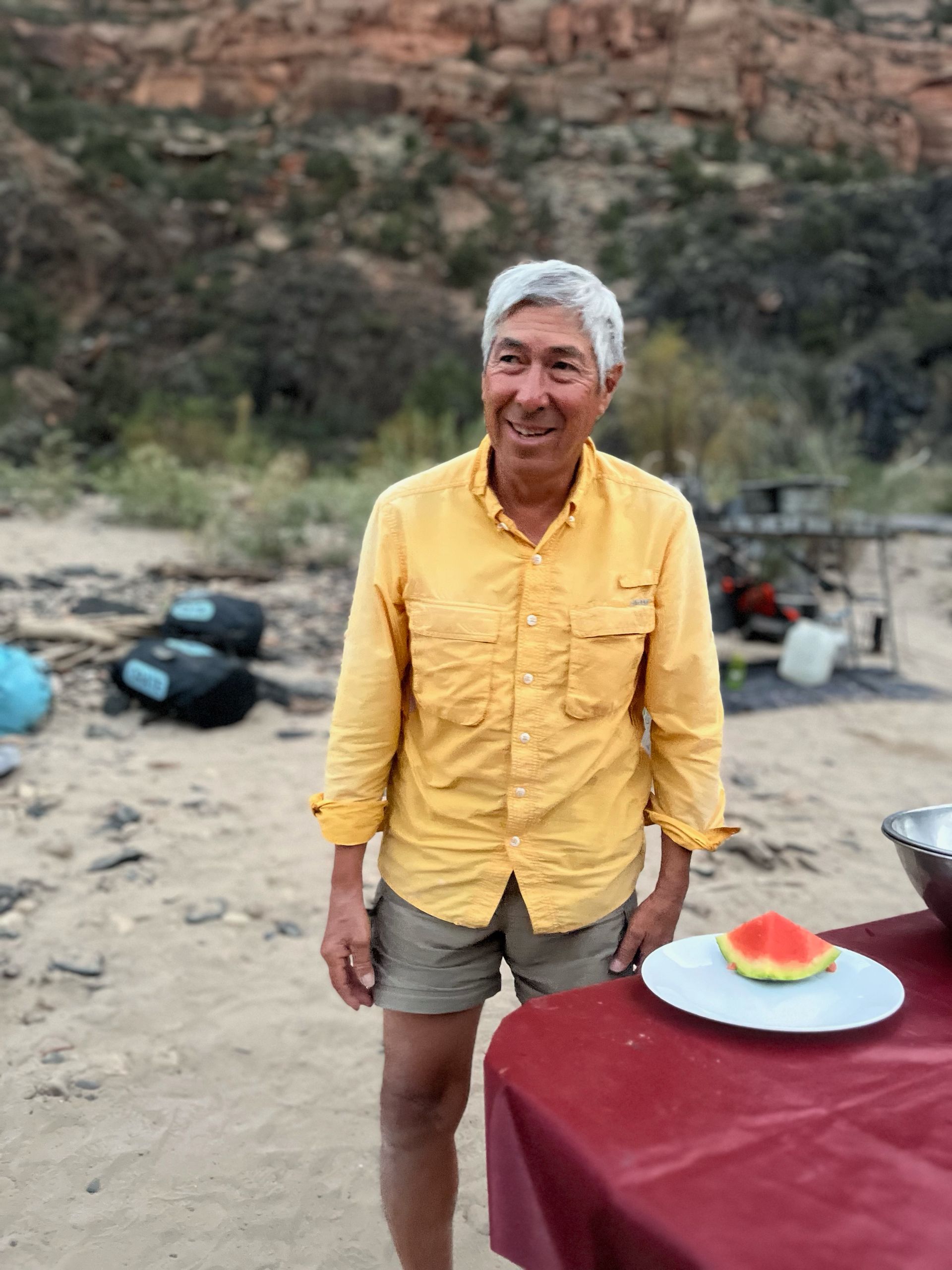 Man in yellow shirt and shorts smiles outdoors near a table with watermelon.