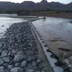 A rocky barrier extends into a river, with mountains in the background.