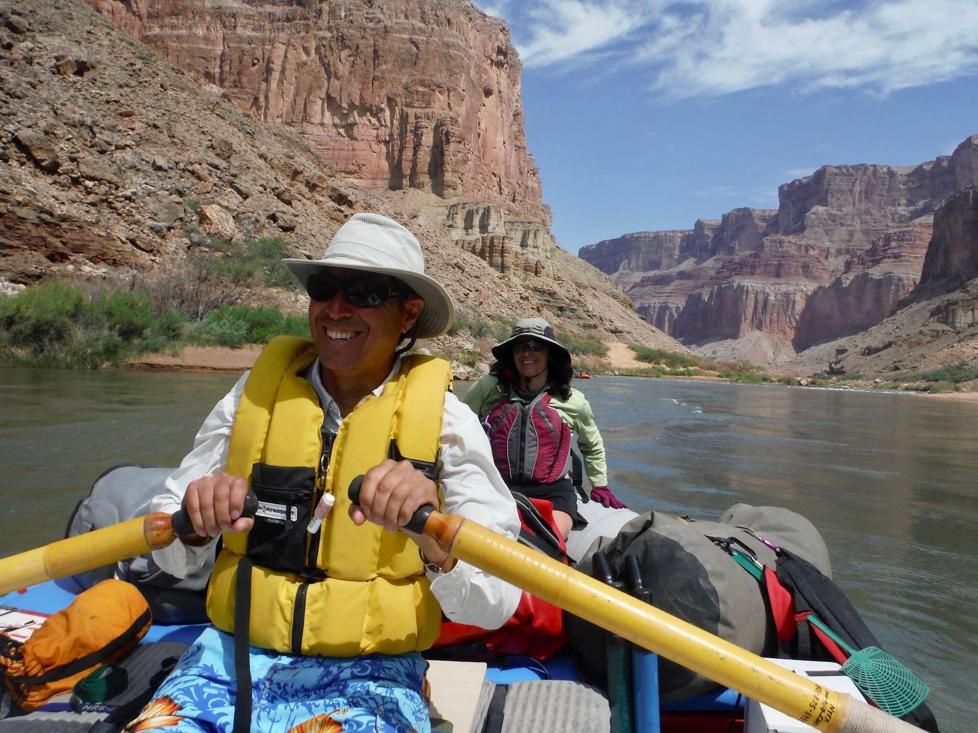 Man rows a raft on a river, with canyon walls in background. Passenger wears a hat. Sunny day.