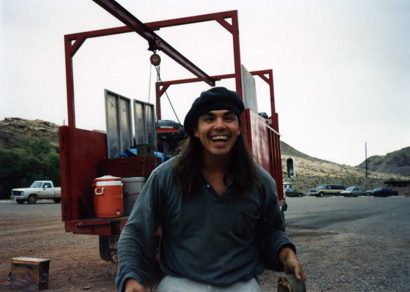 Man with long hair smiles, stands near a red truck in a desert-like setting.