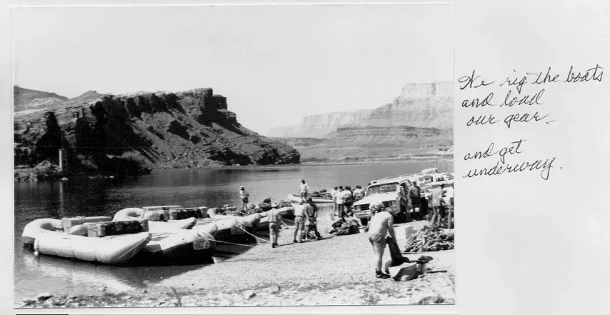 Boats and people on a riverbank, loading gear. Mountain landscape in the background.