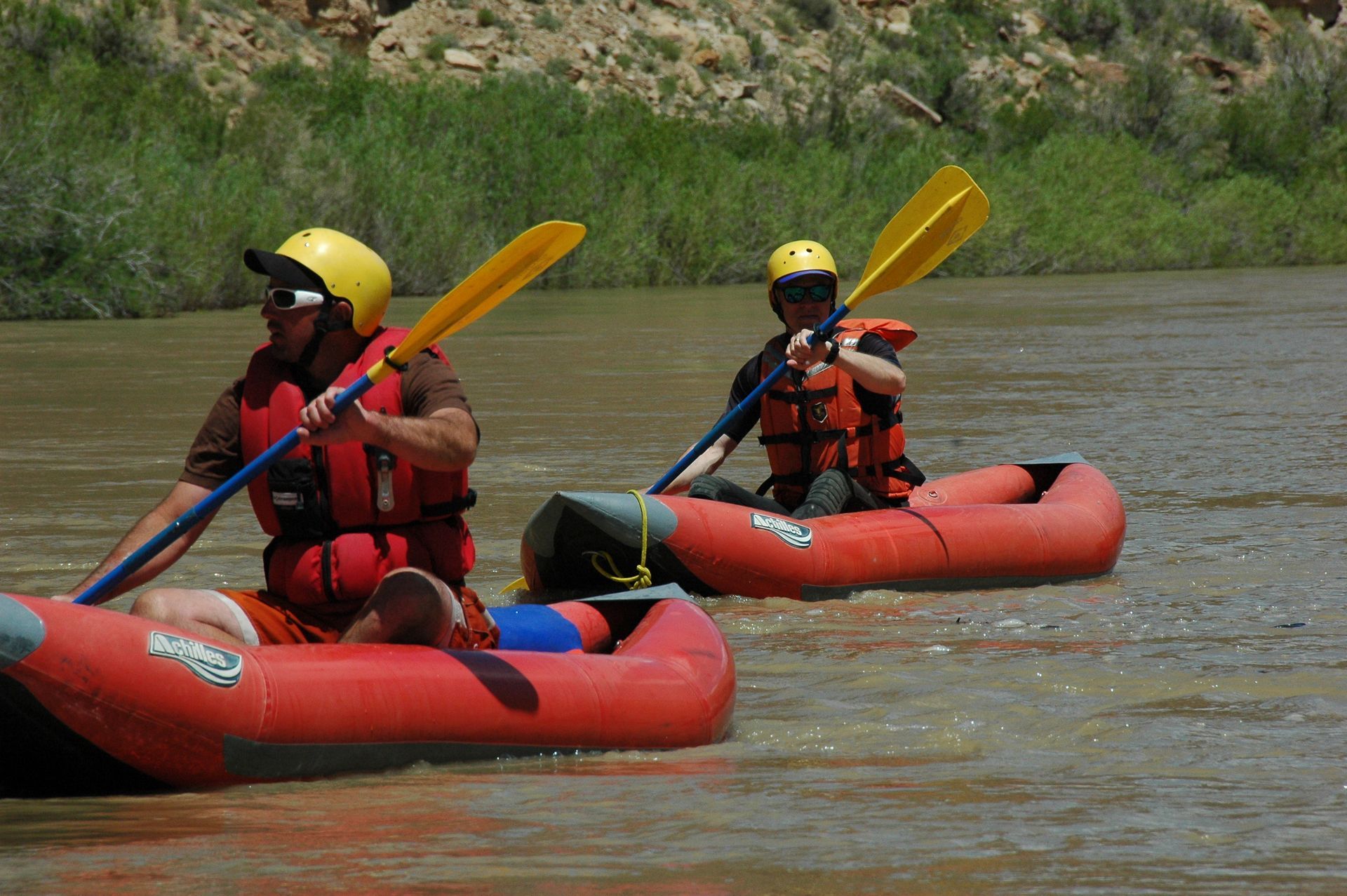 Two people in red inflatable kayaks on a brown river, wearing life vests and yellow helmets.