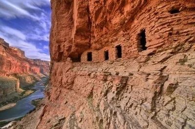 Cliff dwellings carved into a red rock canyon wall overlooking a river.