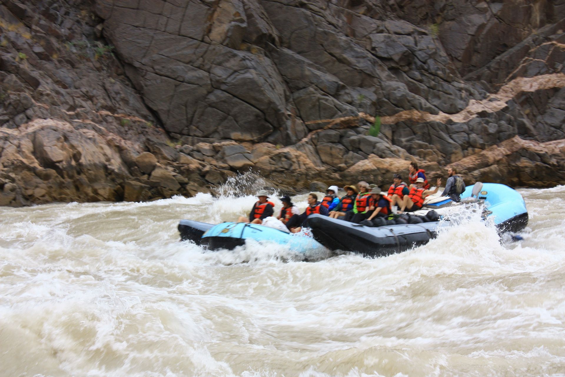 Rafting on a whitewater river; people wear life vests, navigating a rapid near rock cliffs.