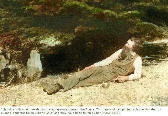 John Muir resting outdoors, hand-tinted photo. Leaning back, long beard, wearing brown overalls, with a cup beside him.
