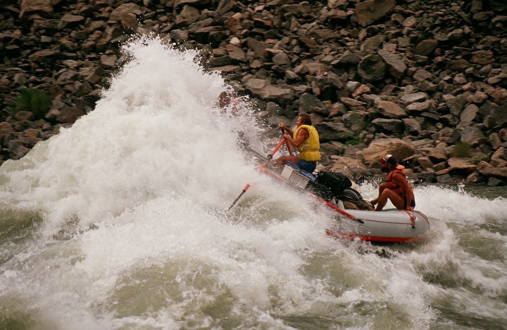 Rafting through whitewater. Two people in raft, one paddling through a large wave. Rocky banks in background.