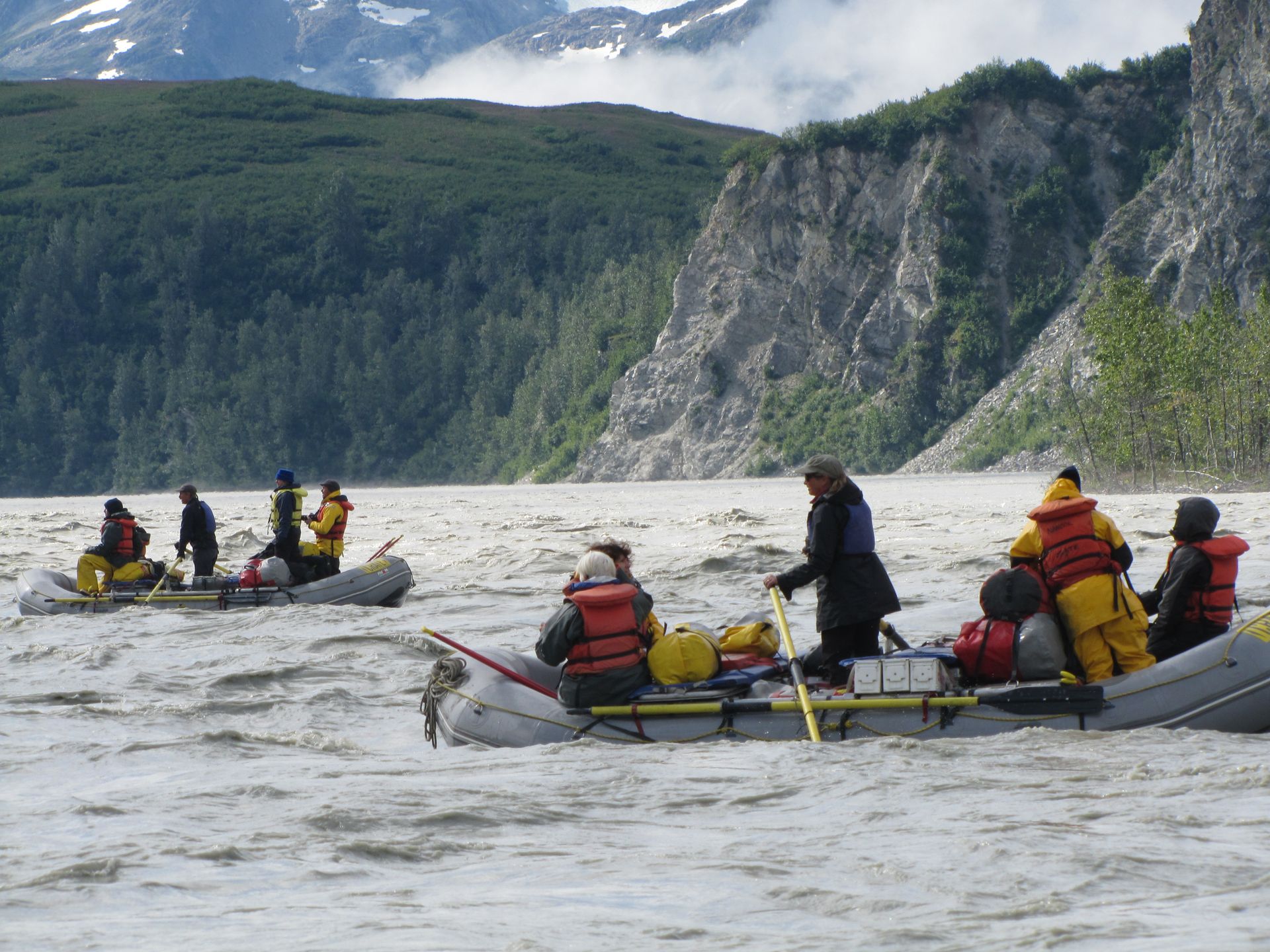 Rafters in inflatable boats navigate a silty river, mountainous landscape in the background.