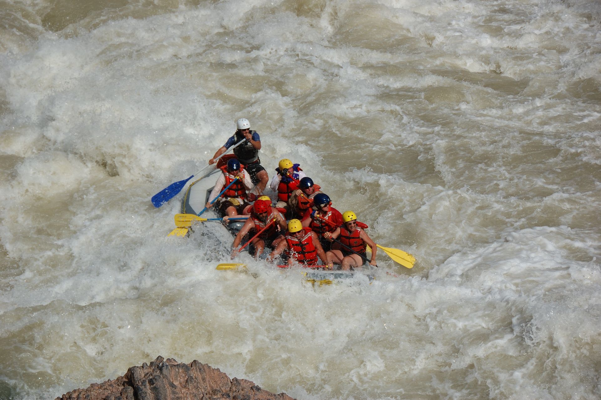 Raft with passengers navigates whitewater rapids, paddlers in red life vests, choppy water.