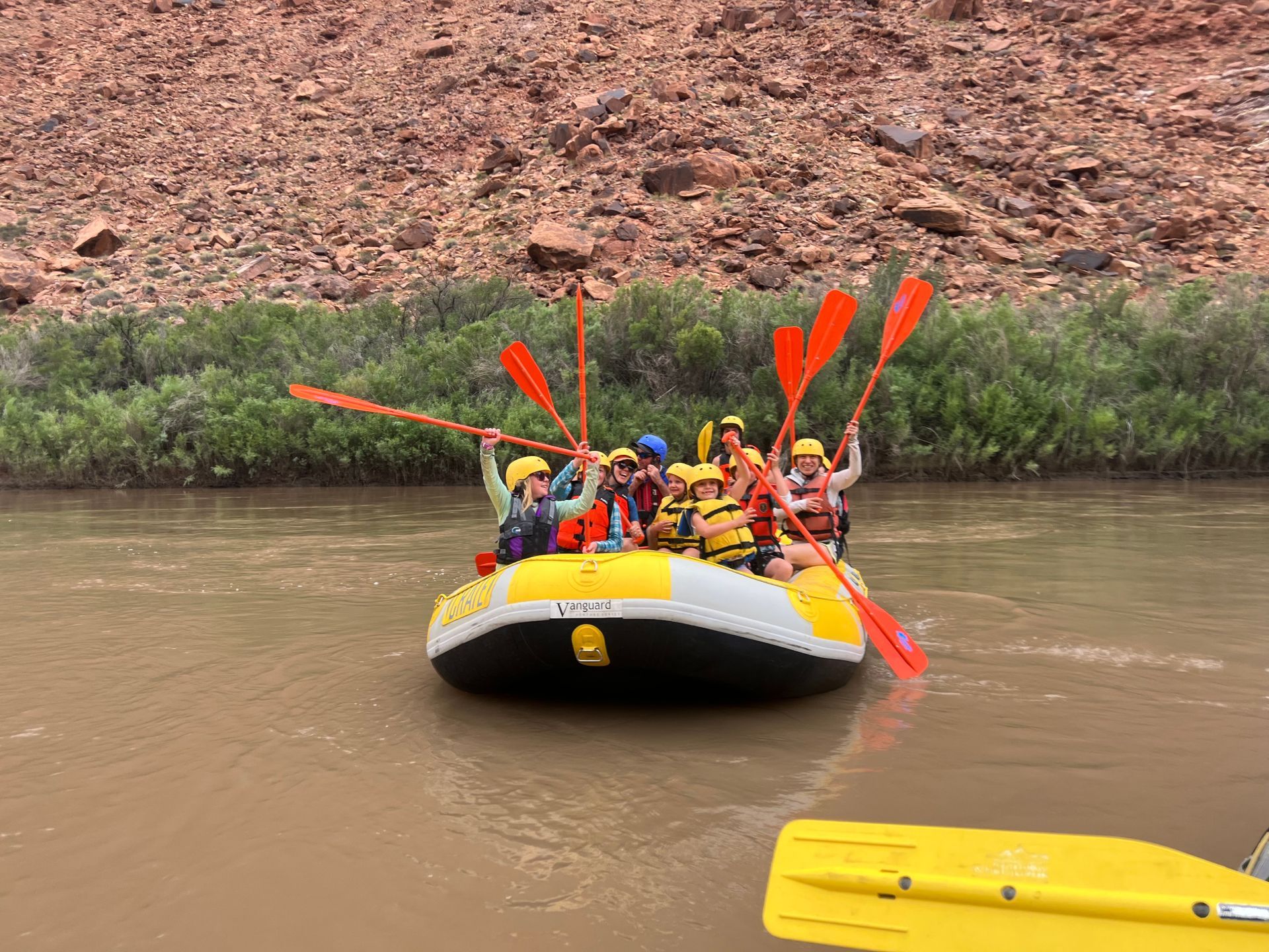 People on a raft with orange paddles, river, red rock backdrop.