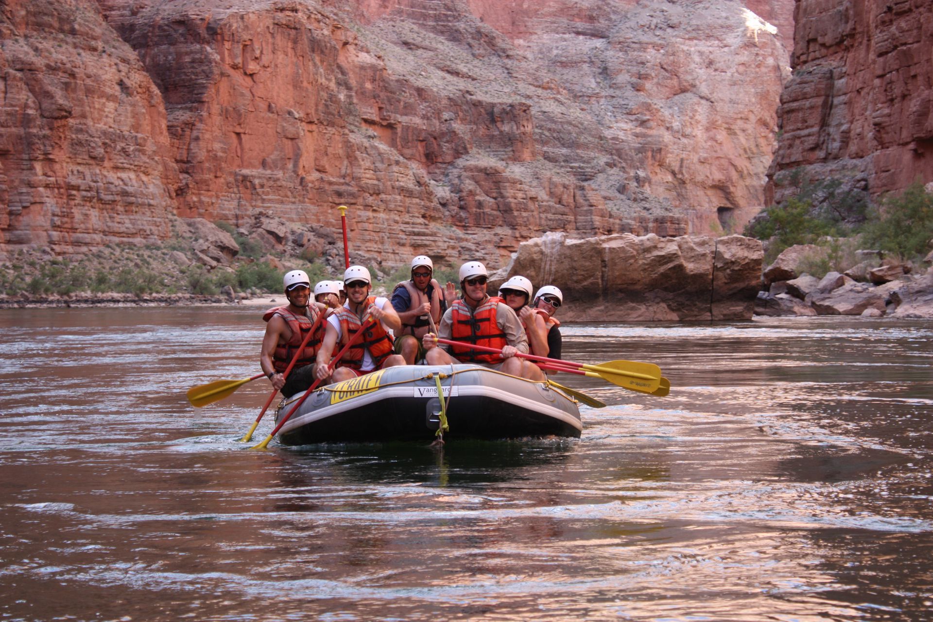 People rafting on a river through a canyon. They are wearing life vests and using paddles.