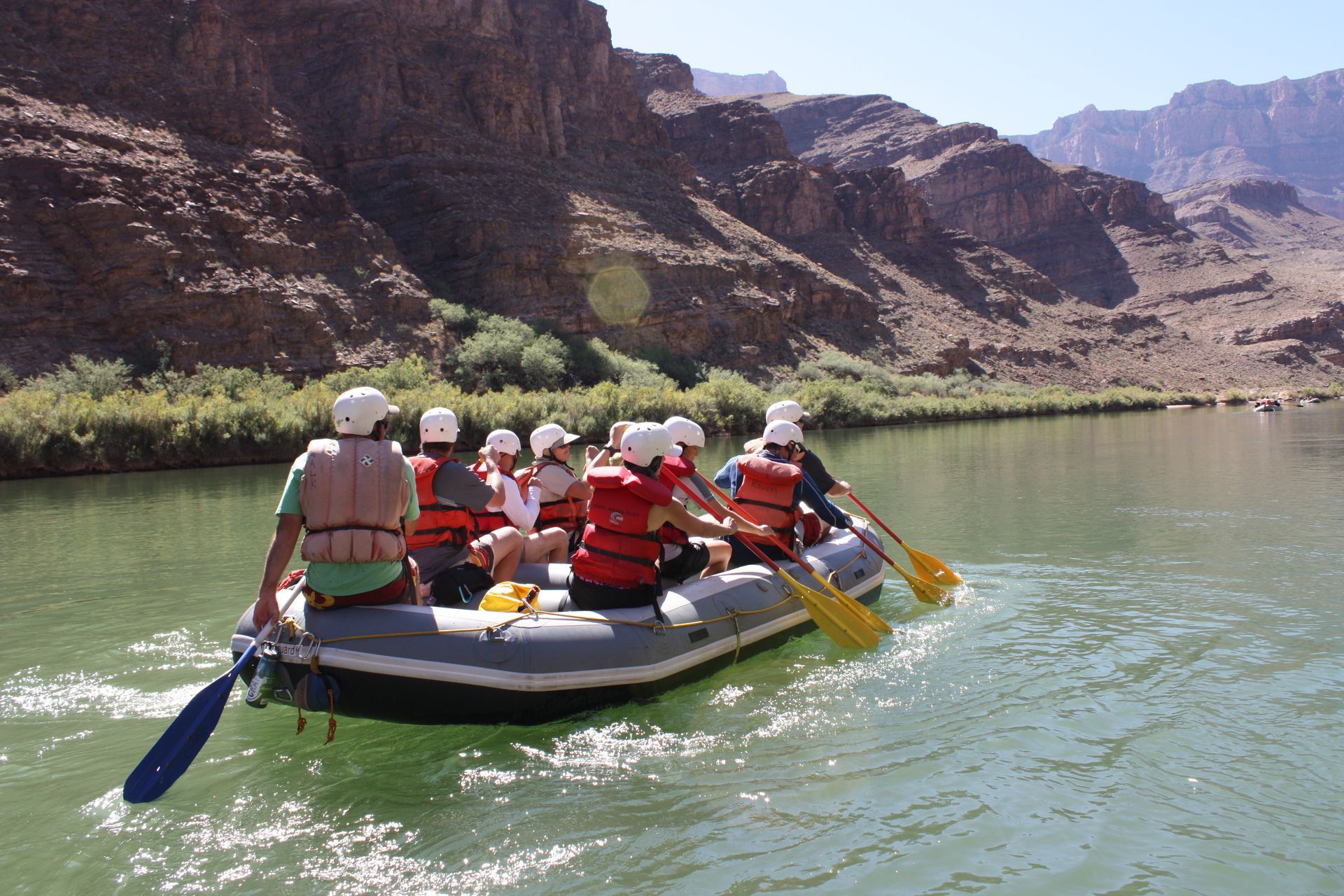 People in a raft with paddles on a river near brown cliffs.