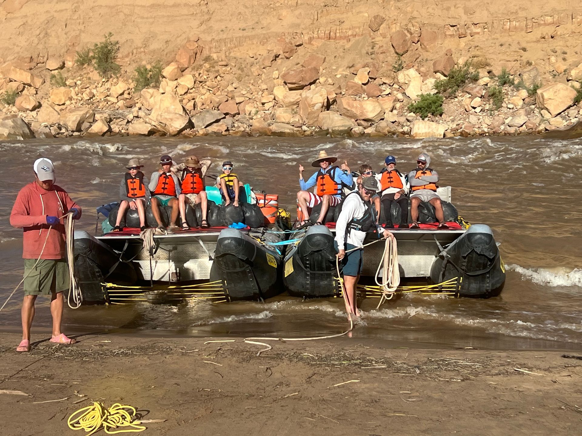 Group on raft in river. People wearing life vests, rocky bank, two guides.