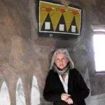 Woman with gray hair leans against a wall, a yellow and black painted sign above.