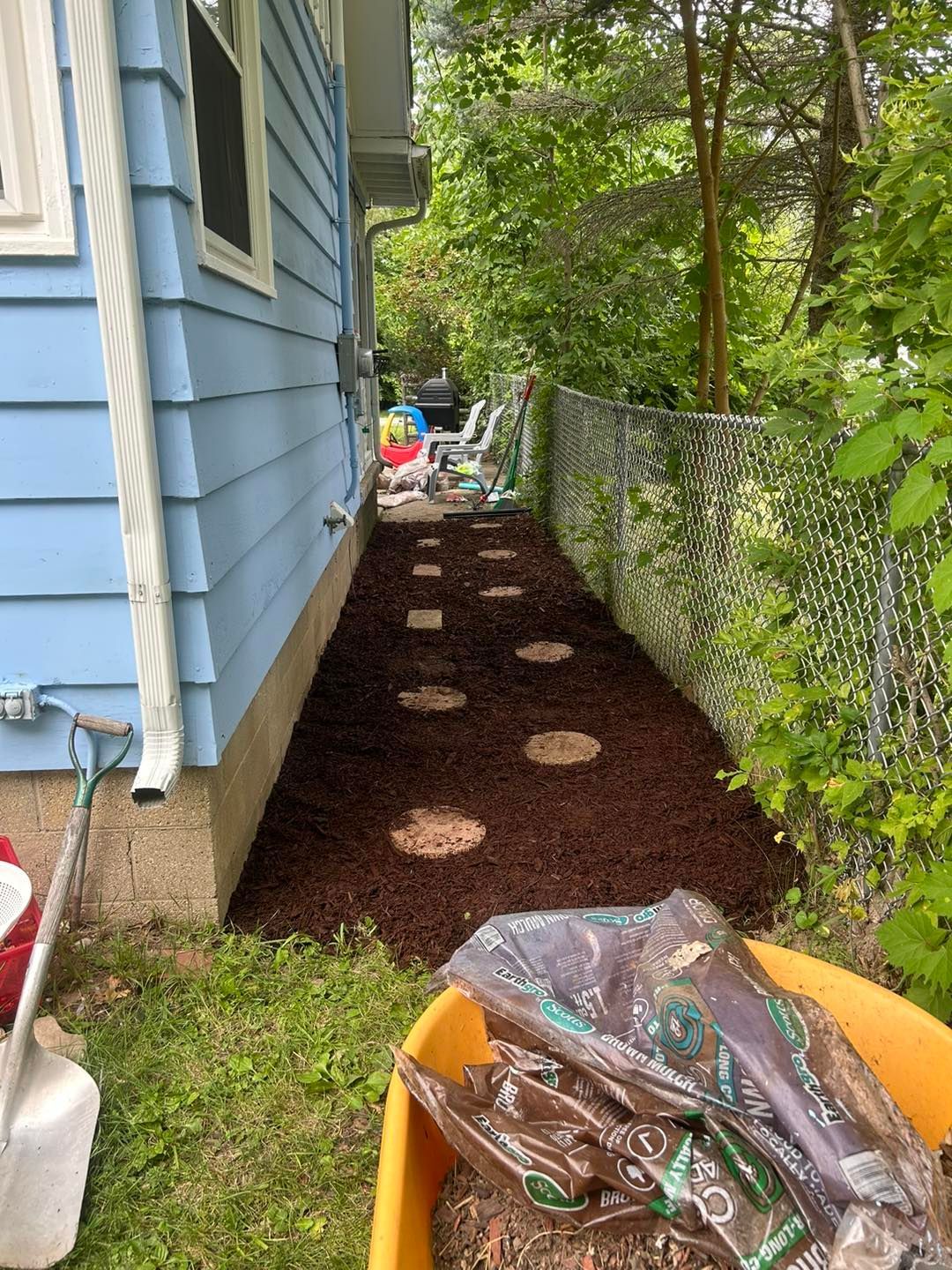 A yellow wheelbarrow filled with mulch next to a blue house.