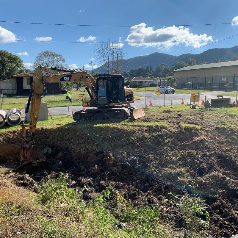 An Excavator Digs Into Dirt on a Grassy Slope Near a Street — B & D Bunt Earthmoving Contractors in Grafton, NSW