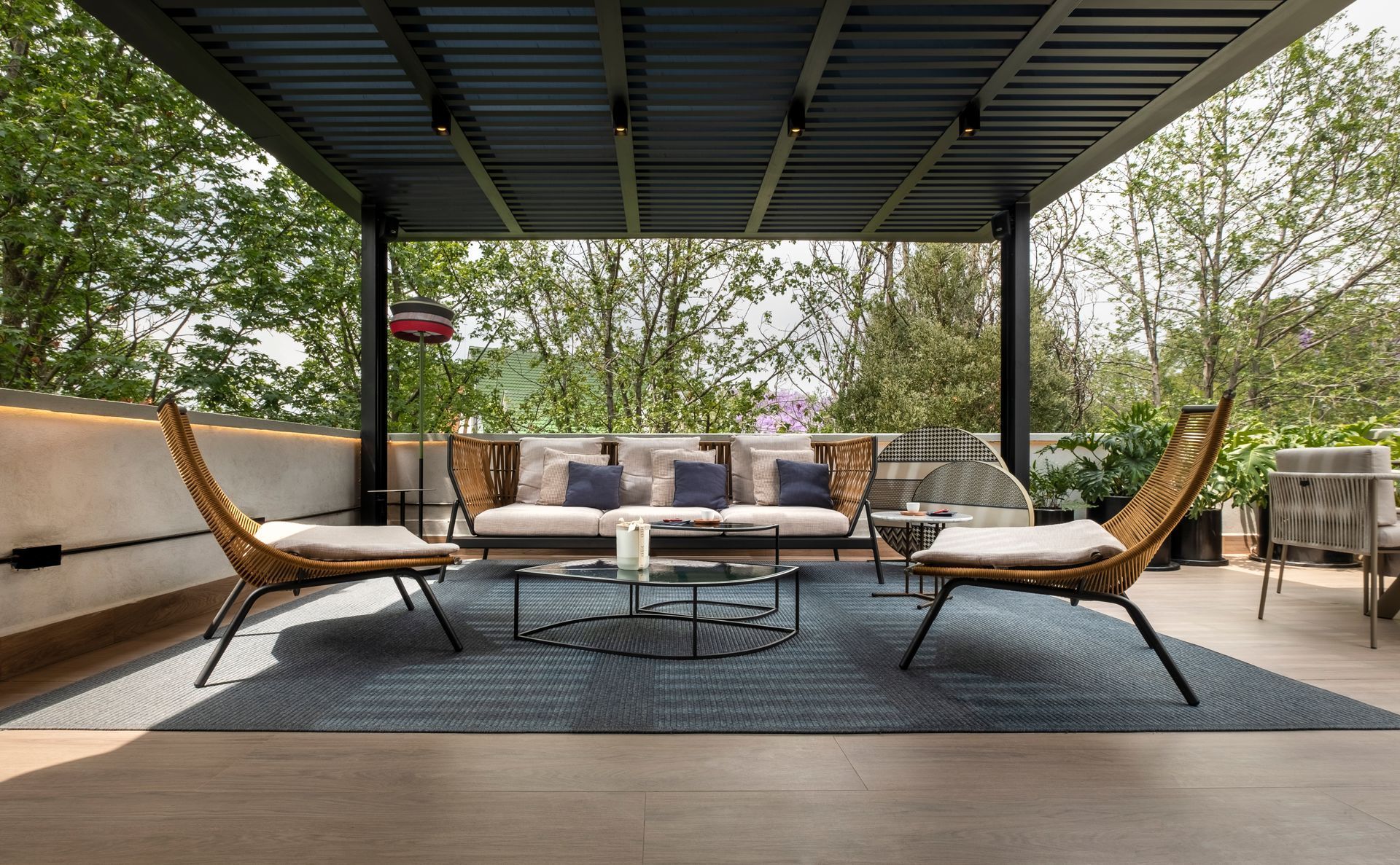 Patio exterior con pérgola, zona de estar, dos sillones, mesa de centro y alfombra, con un frondoso paisaje arbolado de fondo.