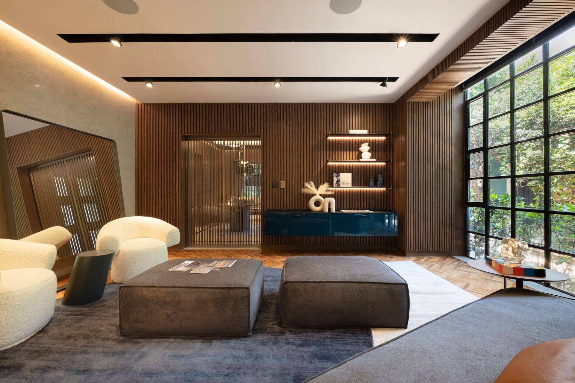 Un salón moderno con paredes revestidas de madera, dos otomanas oscuras, sillas blancas y un gran ventanal que va desde el suelo hasta el techo.