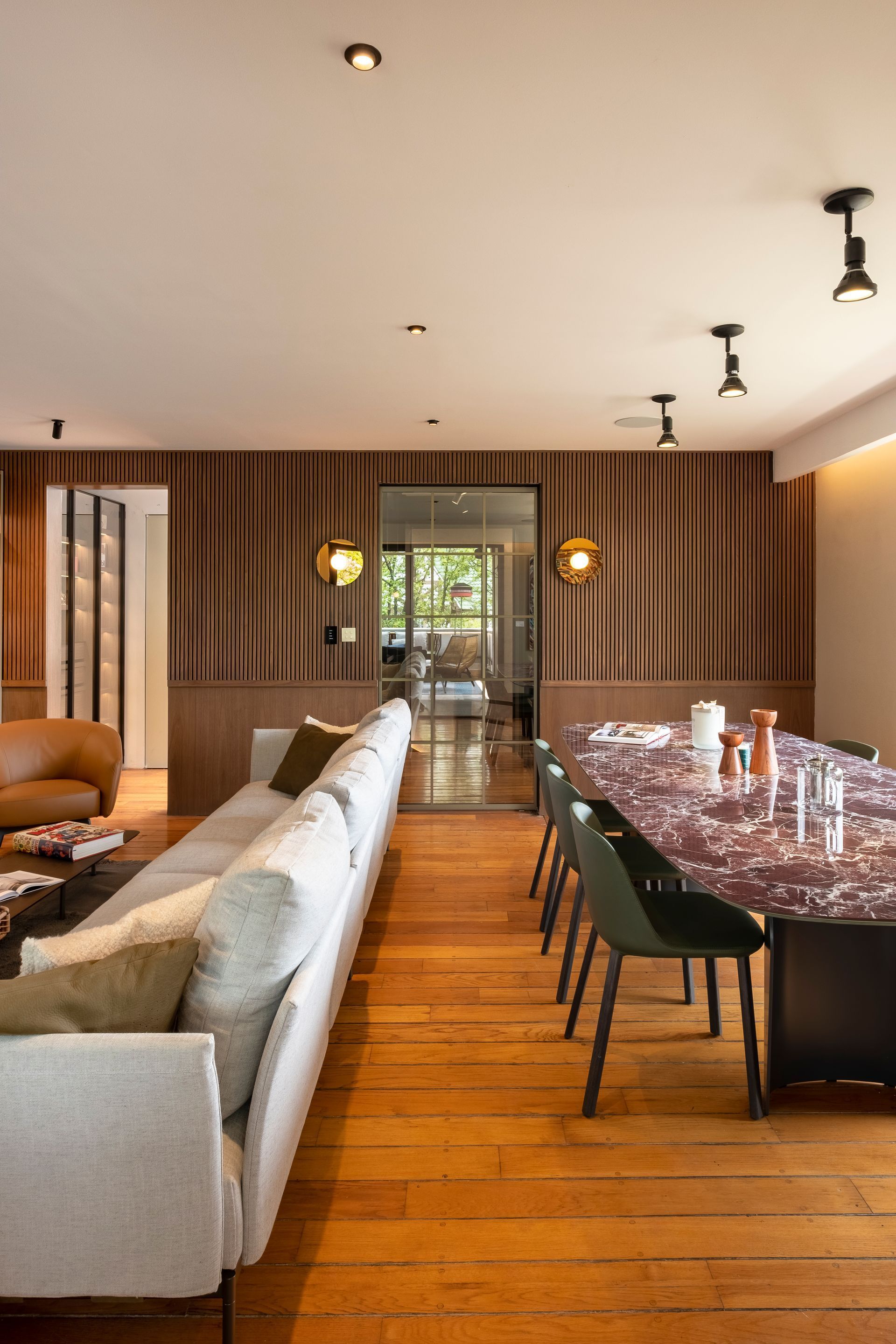 Un moderno salón-comedor con un sofá largo de color claro, mesa de comedor de mármol, paredes revestidas de madera y suelos de madera.