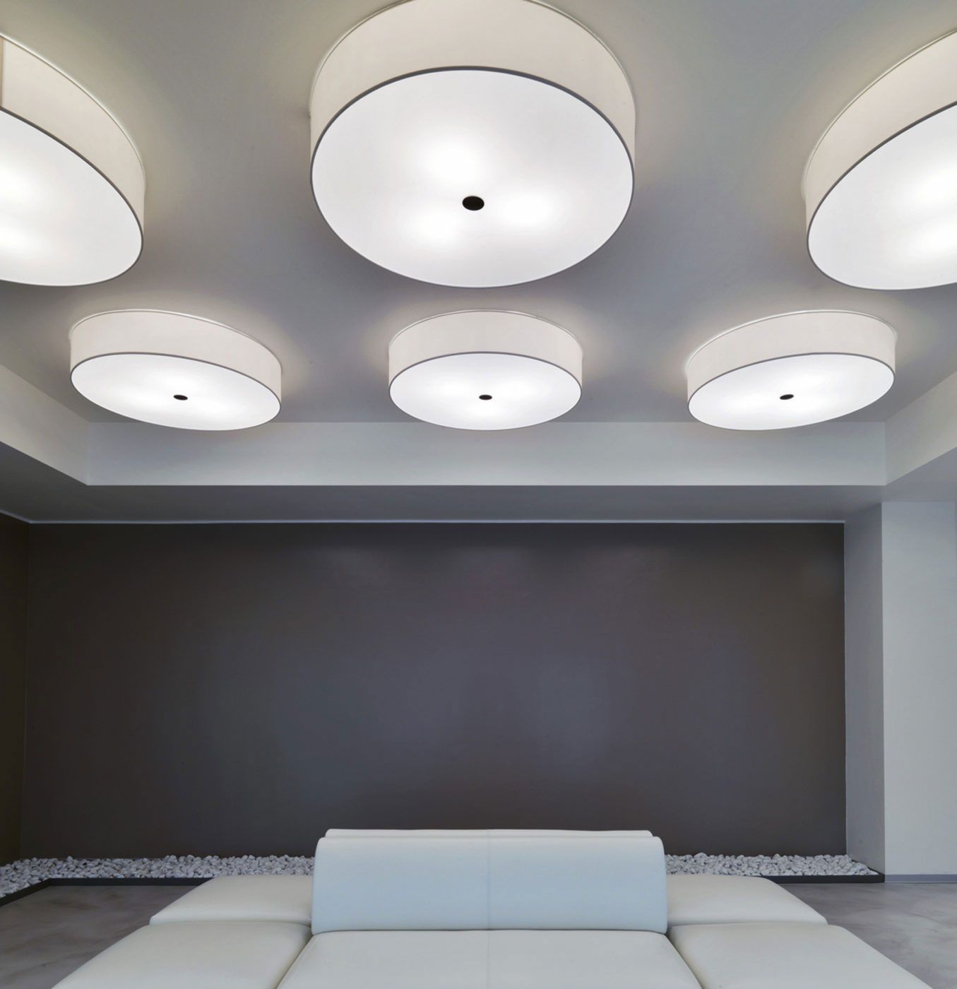 Un salón moderno con un sofá blanco centrado contra una pared oscura, iluminado por seis lámparas de techo circulares y luminosas.