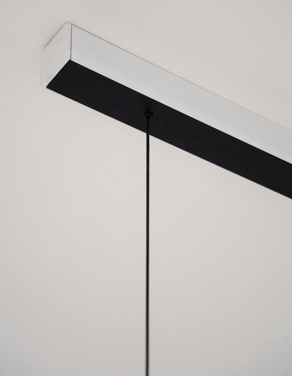 Un fino cordón negro cuelga verticalmente de un riel rectangular blanco y negro en el techo, sobre una pared blanca lisa.