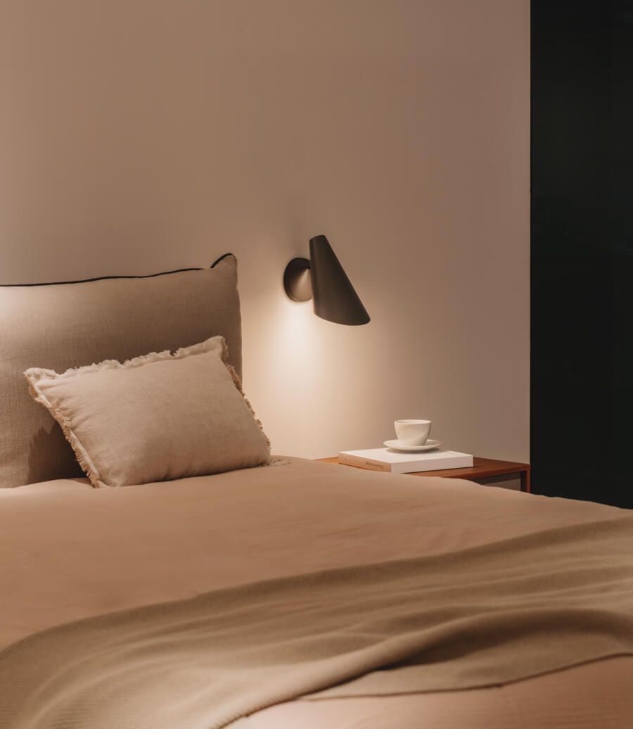 Un dormitorio acogedor y minimalista con ropa de cama beige, una almohada pequeña, una lámpara de pared y una taza sobre una mesita de noche de madera.
