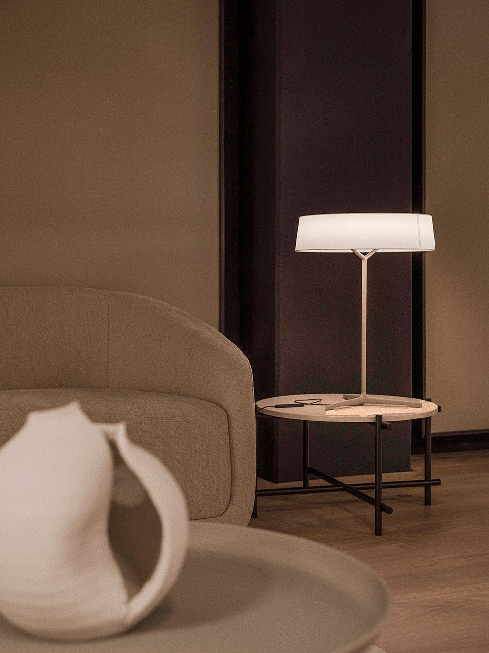 Una habitación minimalista con una lámpara de mesa moderna sobre una mesita auxiliar junto a un sofá texturizado y una escultura abstracta blanca.