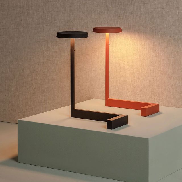 Una lámpara de mesa minimalista en forma de L, de color negro y naranja óxido, descansa sobre una plataforma gris claro contra una pared texturizada.