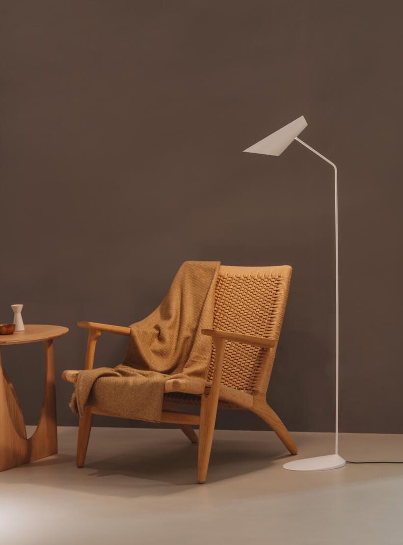Un sillón de madera con respaldo tejido y una manta drapeada se encuentra junto a una pequeña mesa de madera y una lámpara de pie minimalista.