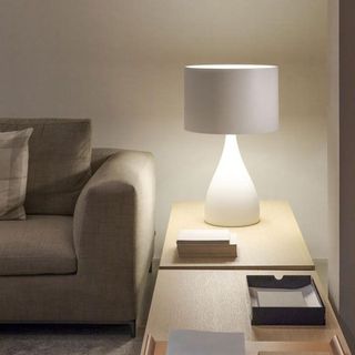 Una lámpara de mesa encendida, con pantalla cilíndrica blanca y base curva, reposa sobre una mesa auxiliar de madera junto a un sofá beige.