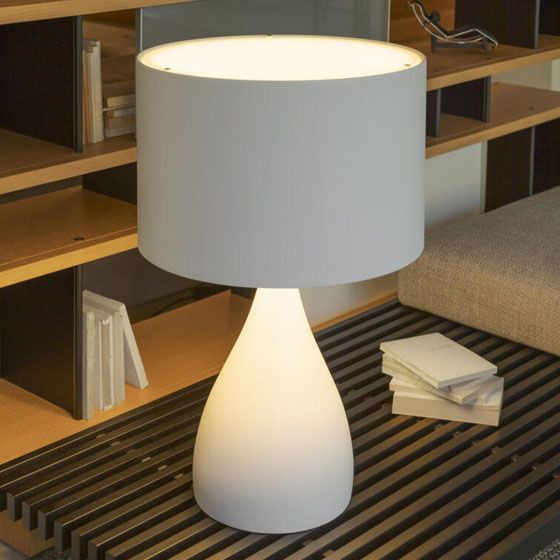 Una moderna lámpara de mesa con una base luminosa en forma de lágrima y una pantalla cilíndrica blanca reposa sobre una superficie de madera con listones.