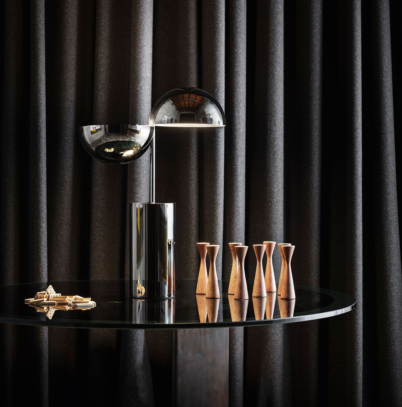 Una moderna lámpara de doble hemisferio con acabado cromado reposa sobre una mesa redonda de cristal con adornos de madera, frente a unas cortinas oscuras.