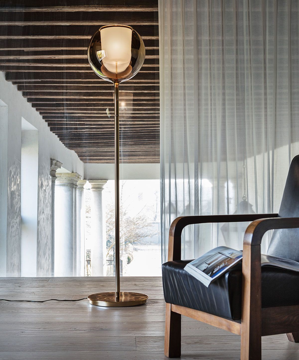 Una lámpara de pie alta con acabado dorado y una esfera de cristal brillante se encuentra junto a un sillón oscuro en una habitación luminosa con techo de madera.