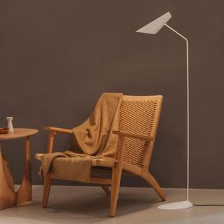 Un sillón de madera clara con respaldo tejido y una manta drapeada se encuentra junto a una mesita y una moderna lámpara de pie blanca.