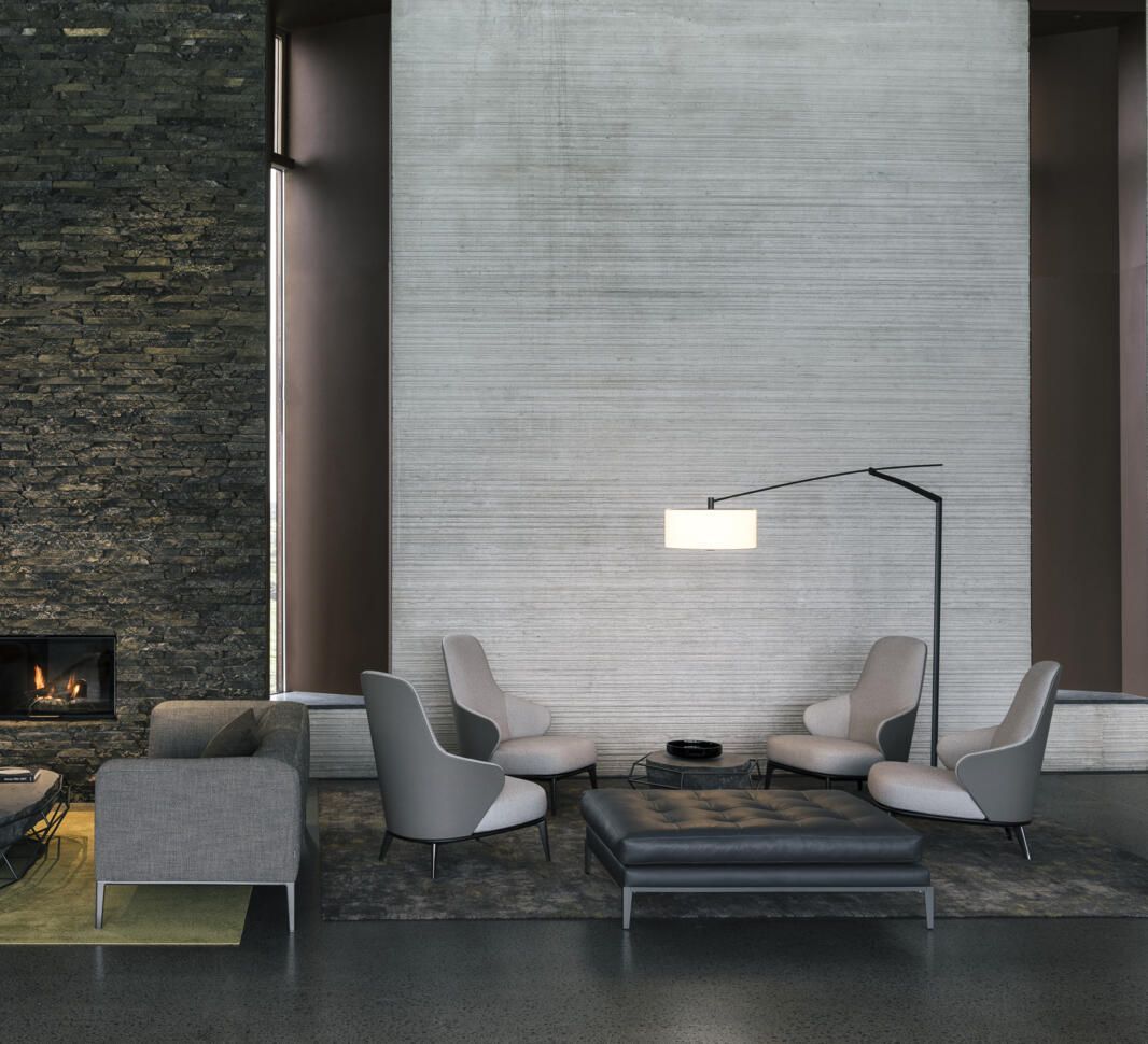 Un salón moderno con sillas grises, un puf de cuero, una lámpara de pie y una chimenea de piedra contra una pared texturizada.