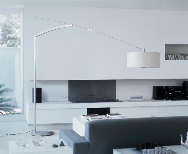 Una lámpara de pie minimalista de color blanco, con brazo arqueado y pantalla cilíndrica, preside un salón moderno decorado en tonos blancos.