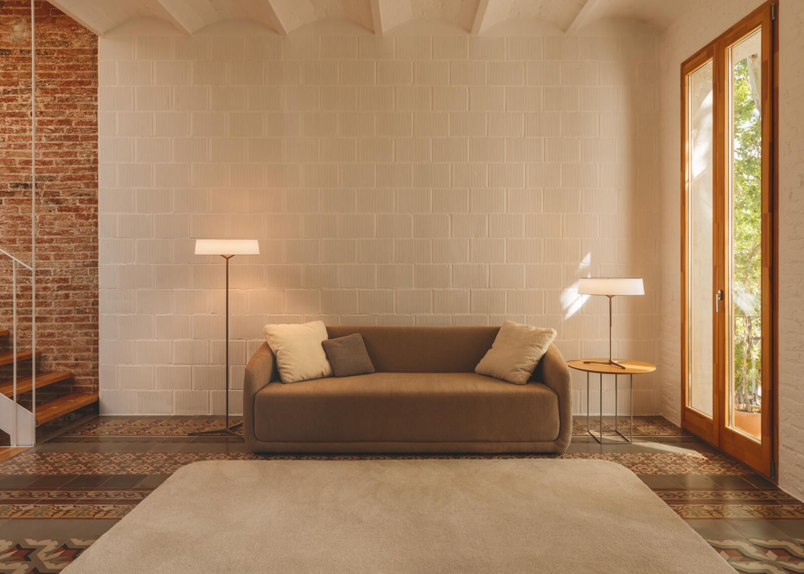 Un salón moderno con un sofá beige, dos lámparas, una alfombra blanca y una pared de ladrillo junto a las escaleras y una ventana.