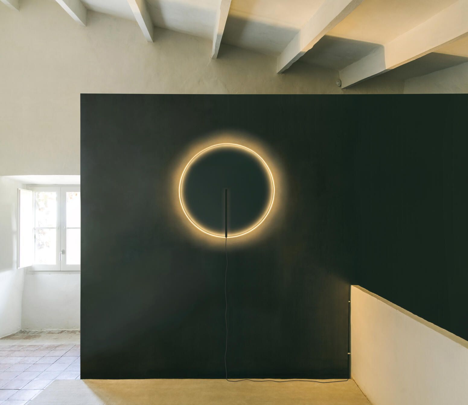 Una luz de neón circular brillante montada en una pared de color gris oscuro en una habitación minimalista y luminosa con vigas blancas.