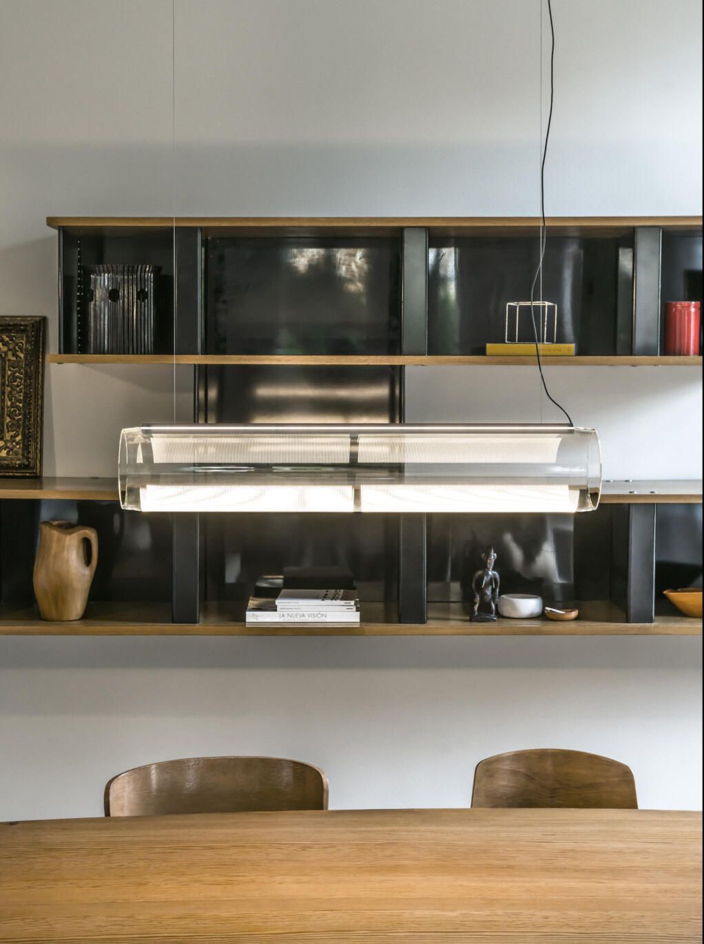Una moderna lámpara colgante horizontal de cristal cuelga sobre una mesa de comedor de madera, frente a una estantería oscura.