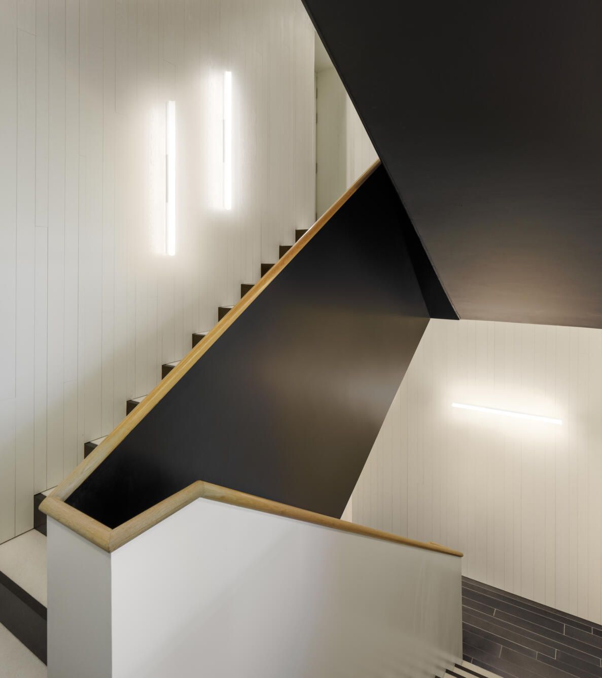 Una escalera moderna con paredes blancas, detalles en negro e iluminación lineal minimalista en la pared.