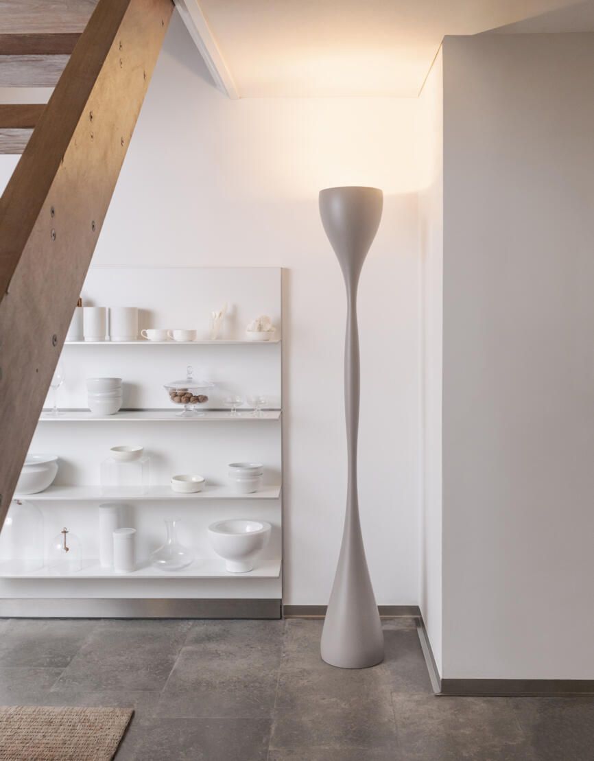 Una lámpara de pie alta y escultural se alza en una habitación moderna junto a estantes blancos repletos de cerámica blanca minimalista.